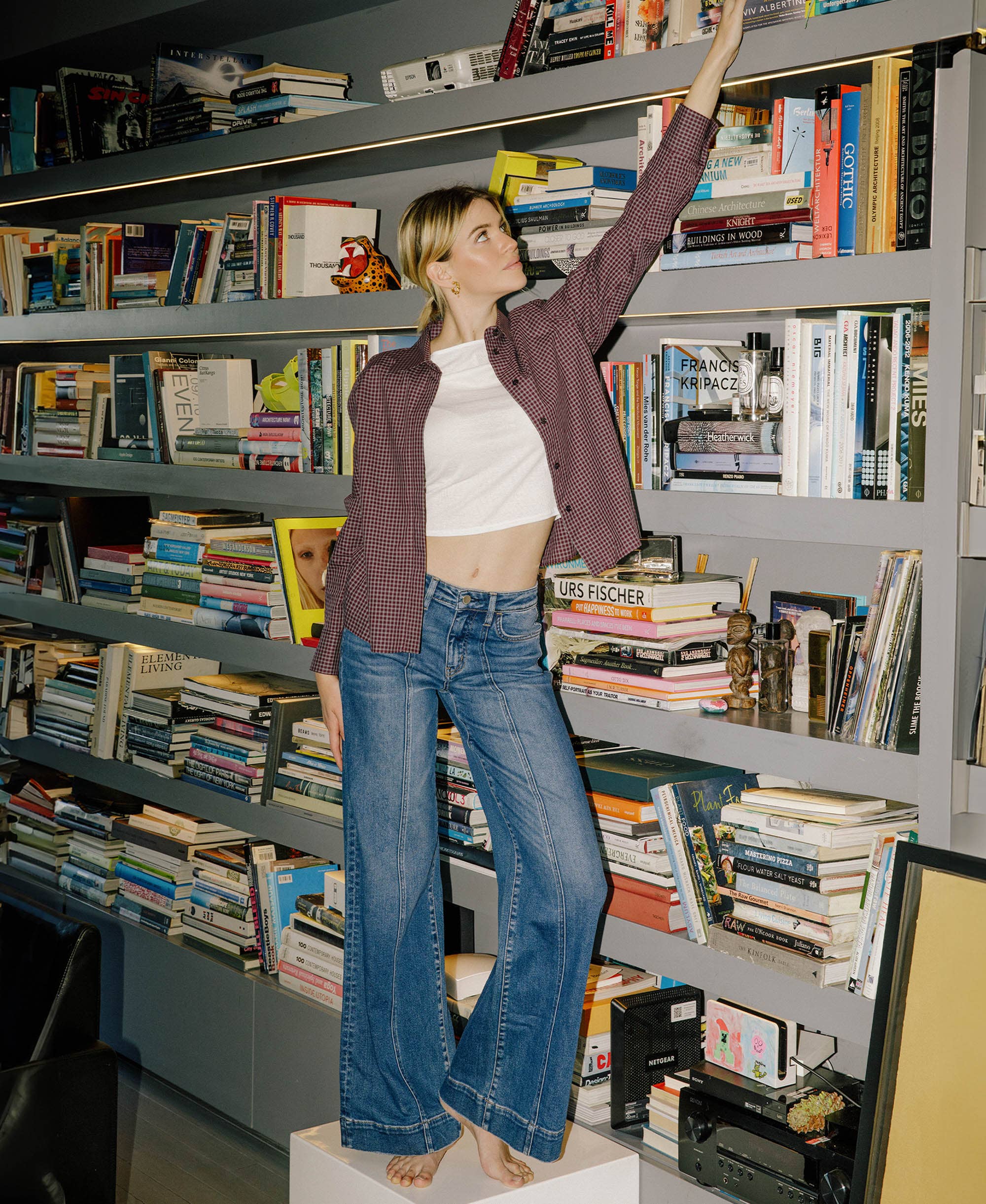 Woman wearing high rise Wide Leg Jeans trouser hem extreme wide leg jeans and a casual shirt standing on a bookshelf in a library setting