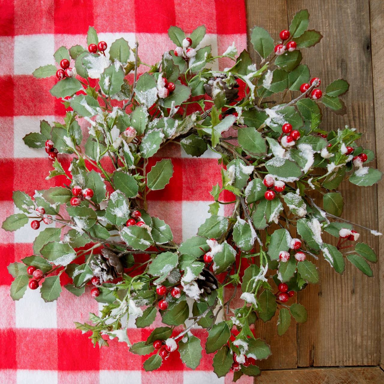 15" glittered holly berry and pinecone wreath with snow on green leaves on red and white checkered cloth background