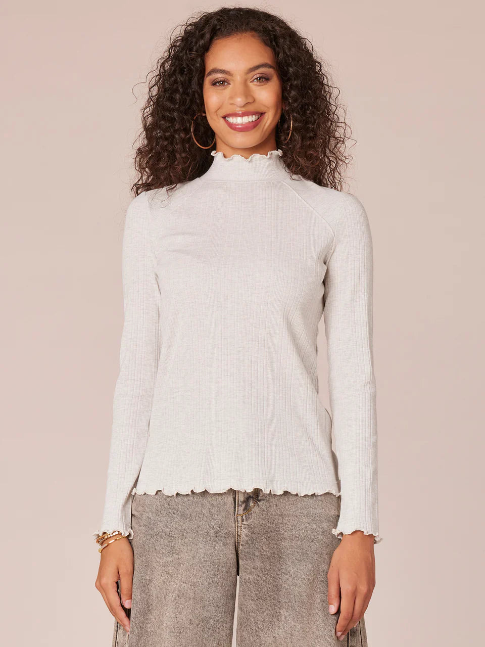 Woman wearing a long raglan sleeve mock neck knit top with lettuce edge in heather cream