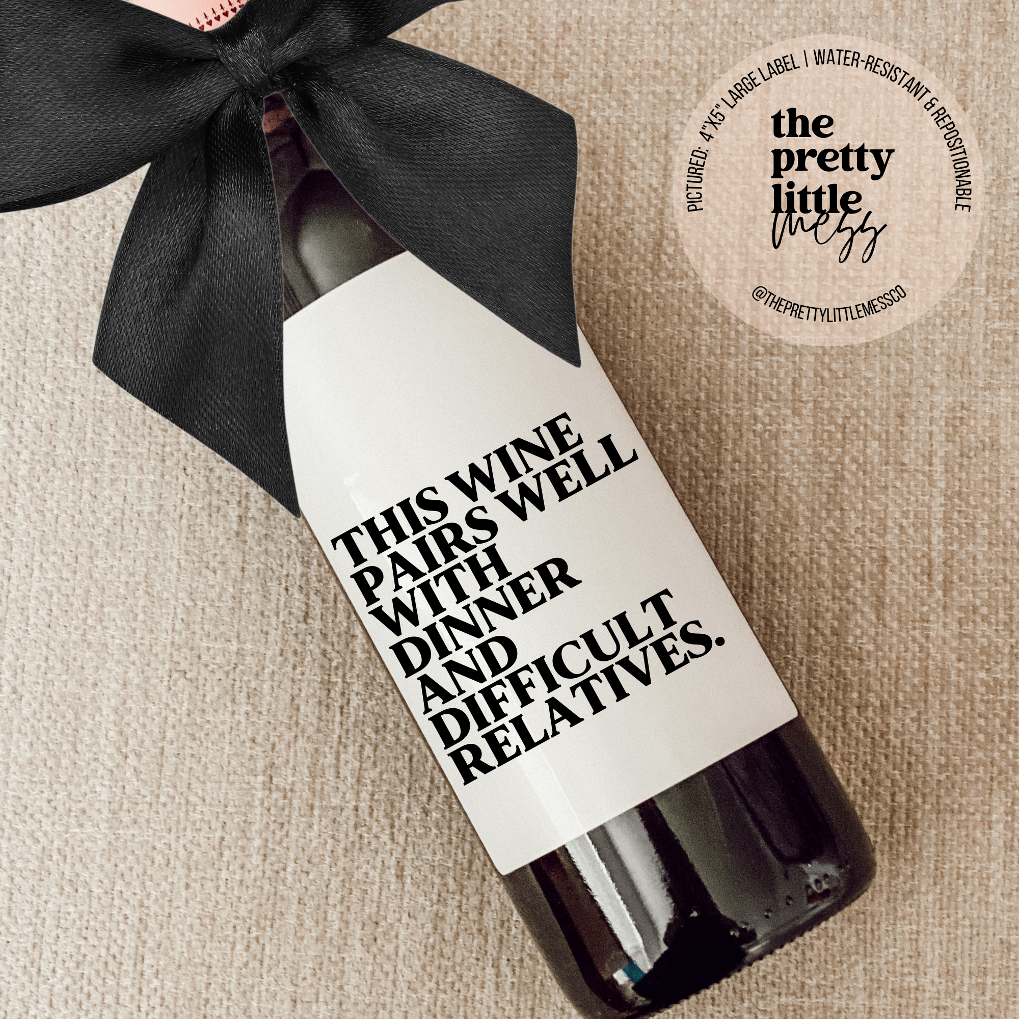 Wine label on bottle reading "This wine pairs well with dinner and difficult relatives" with a black bow on a beige fabric background